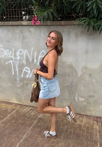 Joven mujer en un top halter y falda de mezclilla posa sobre una pierna, sosteniendo un bolso de flecos marrón con una flor, frente a una pared cubierta de graffiti.
