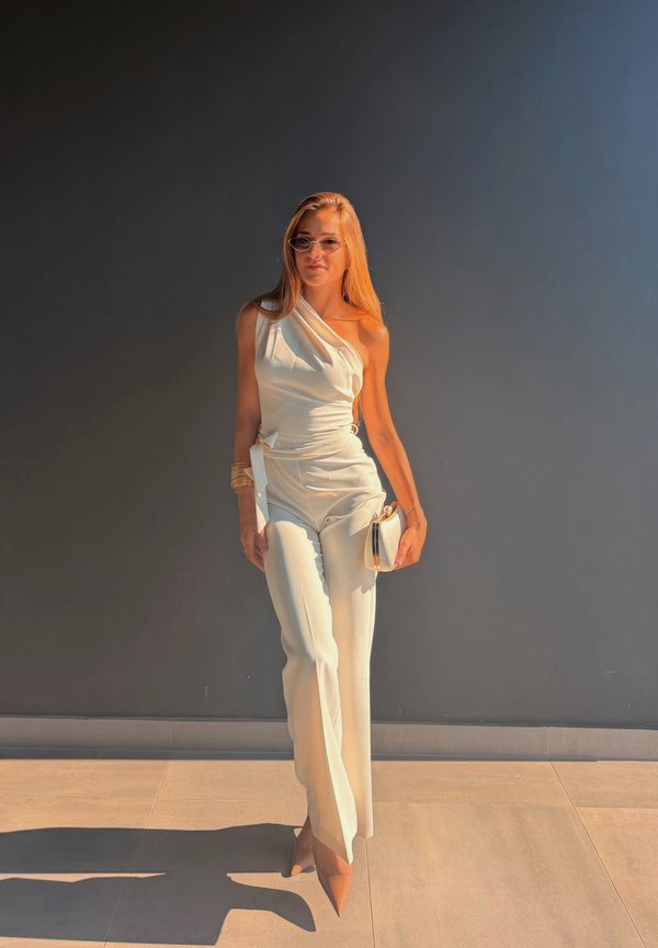 Femme portant une combinaison blanche asymétrique et des talons, tenant une petite pochette blanche, debout contre un mur sombre en plein soleil.