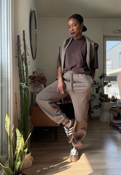 Mujer con atuendo casual marrón y beige está de pie en el interior con una pierna doblada, cerca de un cactus alto y plantas frondosas junto a una ventana con luz natural.