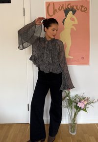 Schwarz-weiß gepunktete Bluse mit Rüschensaum und Glockenärmeln, kombiniert mit hochgeschnittenen schwarzen Schlaghosen, braunen Knöchelstiefeln und Blumen.