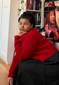 Κόκκινος ριγέ σκούφος με μακριά μανίκια φορεμένος πάνω από ένα μαύρο ρούχο. Το φόντο περιλαμβάνει ράφια γεμάτα βιβλία και μια αφίσα στον τοίχο.