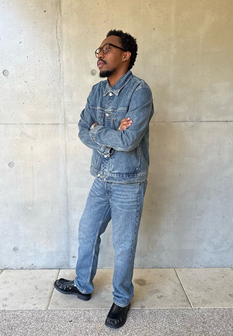 Homme avec des lunettes portant une veste en jean et un jean, debout les bras croisés contre un mur en béton.