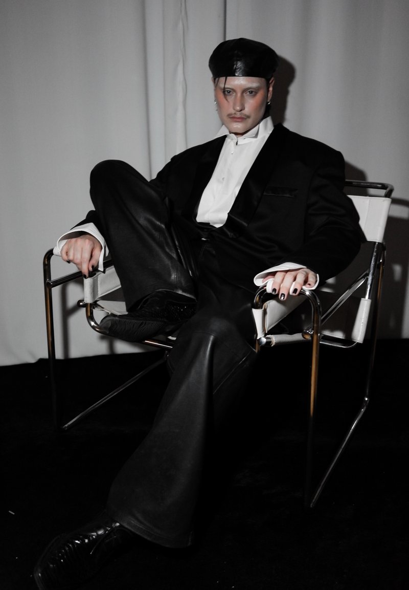 Personne portant un pantalon en cuir noir, une veste noire, une chemise blanche et un béret noir assise avec une jambe relevée sur une chaise à armature métallique sur fond gris.