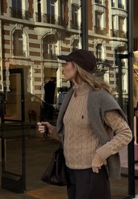 Beige kabelstrikket genser med høy hals, grå sjal drapert over skuldrene, mørk brun lue og svart håndveske. Urban bakgrunn reflektert.