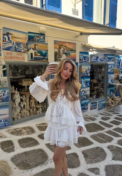 Blusa blanca de capas con volantes y falda corta, fabricada en tejido ligero, con mangas acampanadas. Sosteniendo un vaso de café para llevar, rodeada de exposiciones artísticas.