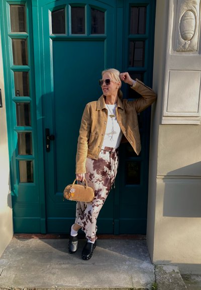 Chaqueta de gamuza marrón, blusa blanca, pantalones marrones con estampado y zuecos negros. Sosteniendo un pequeño bolso de mano marrón. De pie junto a una puerta azul verdosa.