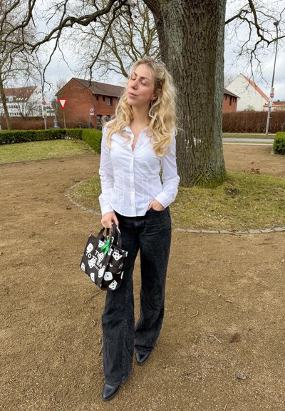 Mujer con cabello rubio largo junto a un árbol grande, vestida con una blusa blanca, pantalones negros de pierna ancha y sosteniendo un bolso de mano negro con estampado de dados.