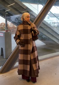 Bruine en beige geruite wollen jas met een hoge kraag, gedragen over een bruine broek, gecompleteerd met zwarte handschoenen en een handtas met patroon.