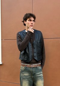 Chaleco de cuero negro sobre una camiseta de manga larga marrón con textura, combinado con jeans azules deslavados y un cinturón estampado. El sujeto tiene el cabello corto y desordenado.