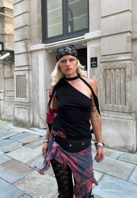 Black asymmetrical top with a choker, layered floral skirt, black lace leggings, and a patterned headscarf. Small red handbag with silver accents.
