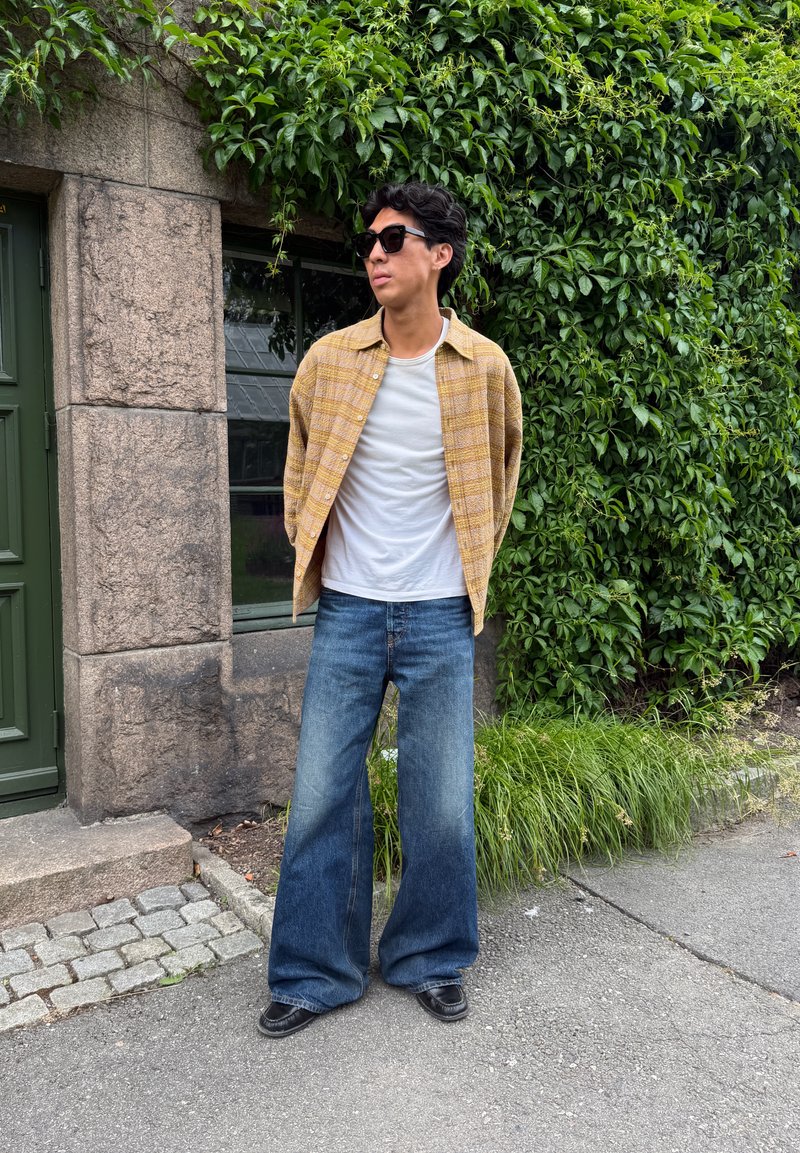 Jeune homme portant des lunettes de soleil, t-shirt blanc, chemise à carreaux jaune, jeans larges et chaussures noires, debout sur le trottoir près d'un mur en pierre et de plantes vertes.