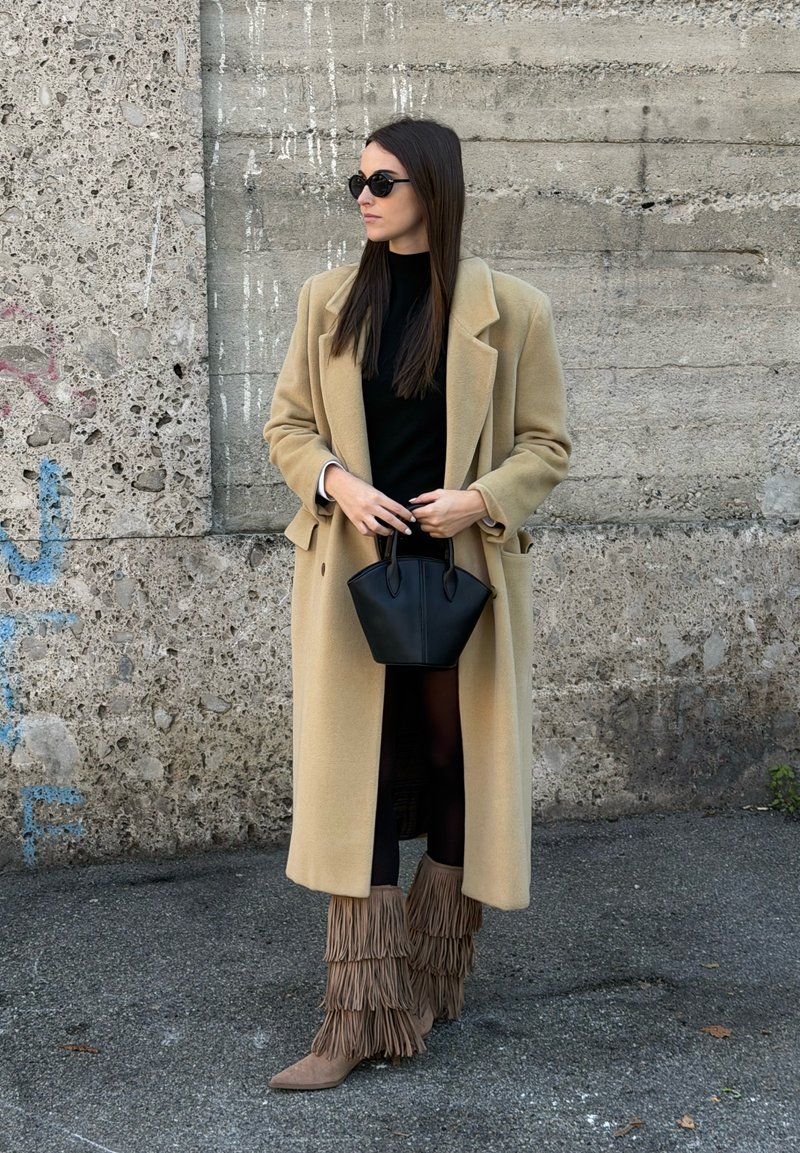 Casaco bege oversize, topo preto, mala preta, botas bege com franjas, óculos de sol, com um fundo de parede cinza texturizada.