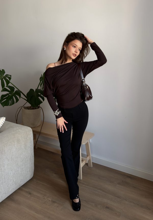 Nő sötét váll nélküli toppban és fekete nadrágban pózol beltéren, növény és pad közelében, egyik kezével a hajában, válltáskát viselve.