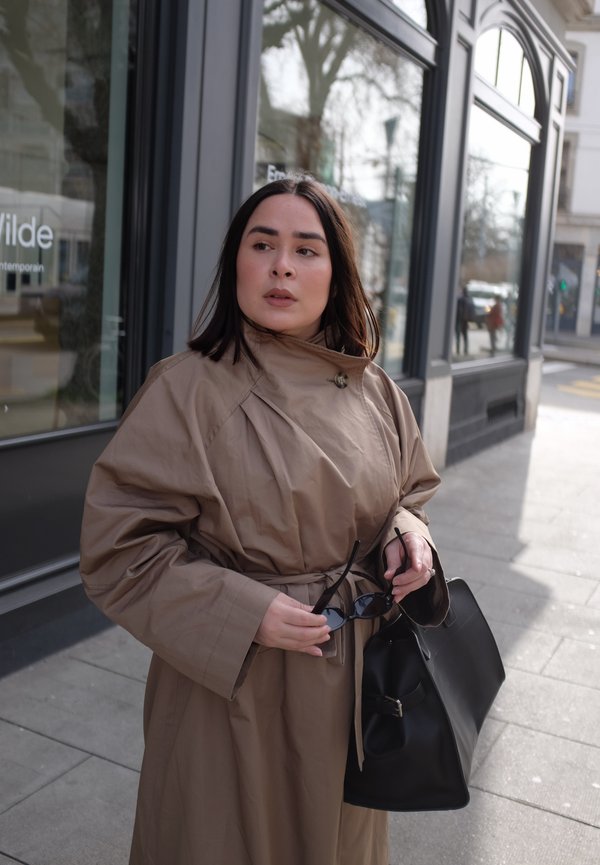 Frau in einem beigefarbenen Trenchcoat hält eine schwarze Sonnenbrille und eine große schwarze Handtasche auf einem Bürgersteig in der Stadt nahe verglasten Ladengeschäften.