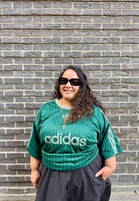 Persona sonriendo con cabello largo y rizado, usando gafas de sol negras, camiseta verde de Adidas, collar de cruz de oro y pantalones negros, de pie contra una pared de ladrillos grises.