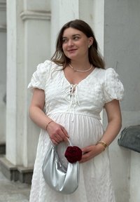 Kvinna i vit texturerad klänning som håller en silverhandväska med röd blomsternot, bärande pärlhalsband och guldarmband, stående nära en vit vägg.