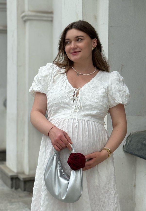 Femme dans une robe texturée blanche tenant un sac à main argenté avec un détail de fleur rouge, portant un collier de perles et une montre en or, debout près d'un mur blanc.