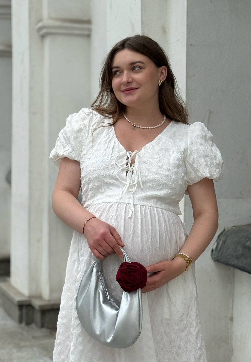Frau in einem weißen strukturierten Kleid, die eine silberne Handtasche mit roten Blumendetails hält. Sie trägt eine Perlenkette und eine goldene Uhr und steht nahe einer weißen Wand.