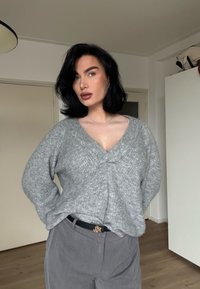 Pull gris en maille avec un col en V, présentant une finition texturée. Associé à un pantalon taille haute gris clair et une ceinture noire.