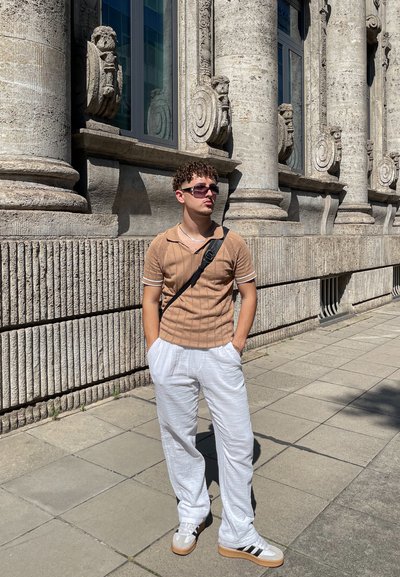 Polo de manga corta tan con tejido texturizado y ribete blanco. Pantalones de lino blancos holgados. Zapatillas grises con rayas negras. De pie frente a una pared de piedra.