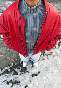 Personne portant une veste rouge par-dessus un pull gris à motifs d'étoiles, un jean bleu clair et des baskets blanches, debout sur un sol enneigé et glacé.