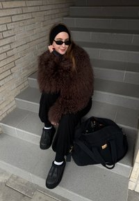 Veste en fourrure brune et touffue, pantalon noir, chaussures noires sans lacets. Sac fourre-tout noir à côté. Escaliers carrelés de couleur claire et mur en pierre texturée en arrière-plan.