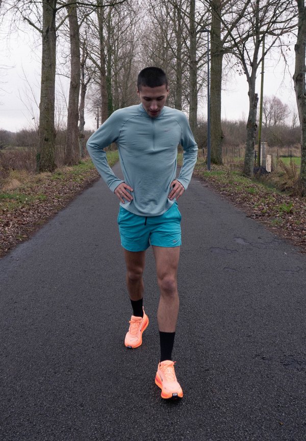 Jeune homme en tenue de sport bleue et baskets néon s'étirant sur une route bordée de feuilles, entourée d'arbres dénudés par un temps couvert.