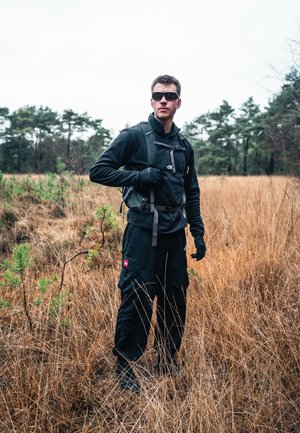 Chaqueta y pantalones negros de outdoor, guantes y gafas de sol; equipado con una mochila estilo bandolera; situado en un campo de hierba con árboles al fondo.