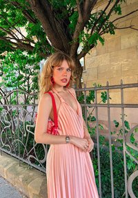 Vestito plissettato rosa pallido con scollatura profonda. Abbinato a una borsa rossa, un orologio argentato e grandi orecchini rossi. Foliage verde sullo sfondo.