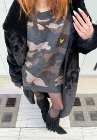 Sivý pletený sveter s maskáčovým vzorom a svetlohnedými akcentmi, skombinovaný s čiernym kabátom z umelého kožušiny a čiernymi členkovými čižmami s trsťami.