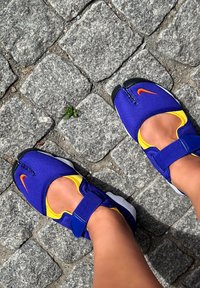 Blue and yellow slip-on shoes with an orange Nike logo, featuring a zip closure and a textured rubber sole, set against a cobblestone background.