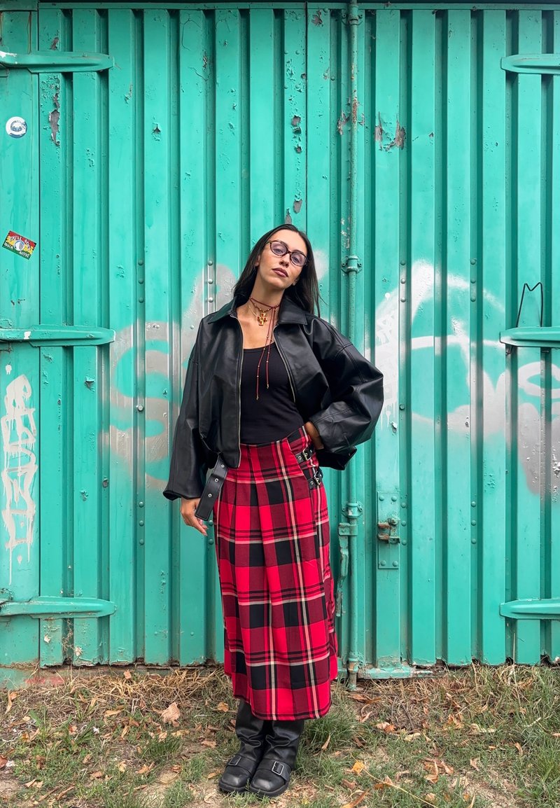 Casaco cropped de couro preto, blusa preta ajustada, saia midi xadrez vermelha, botas pretas até ao joelho, óculos redondos, de pé contra uma parede turquesa.