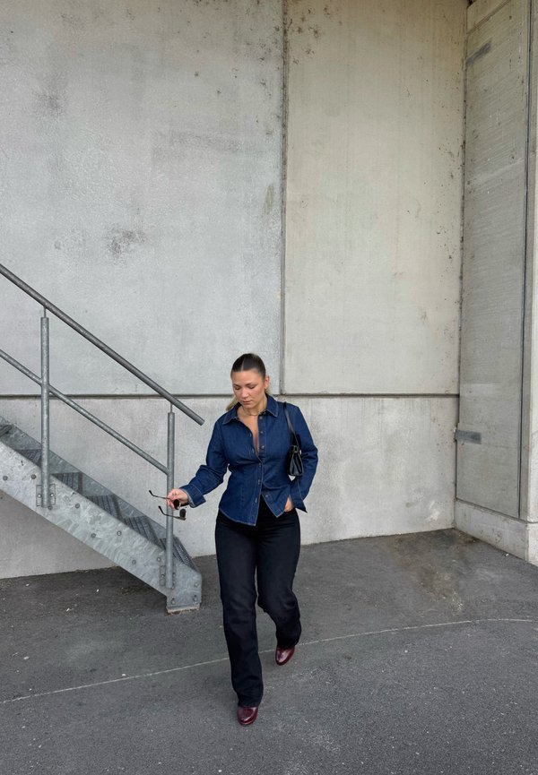 Mujer con camisa vaquera y pantalones negros caminando cerca de unas escaleras de metal, sosteniendo gafas de sol en una mano y un bolso colgado del hombro.