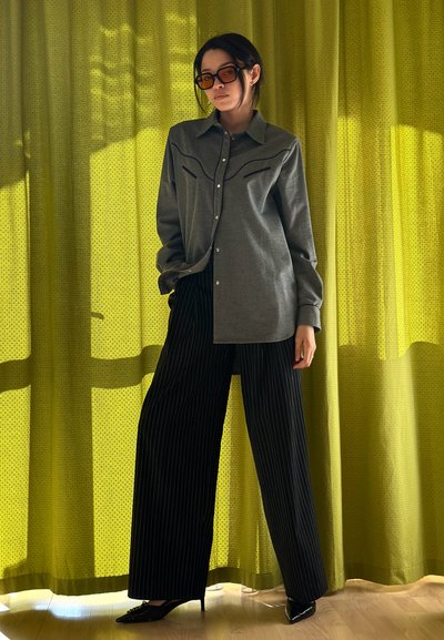 Camisa gris de botones con yugo de estilo occidental y pantalones de pierna ancha a rayas negras, complementados con gafas de sol marrones de gran tamaño contra un fondo verde texturizado.