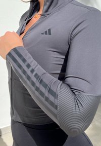 Grå atletisk zip-up jacka med ribbad textur och svarta randiga detaljer på ärmarna. Har en logotyp på den övre vänstra bröstområdet.