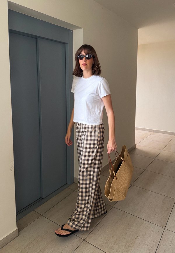 Vrouw met wit T-shirt, geruite broek, zwarte zonnebril, draagt grote stro draagt tas, staand voor gesloten blauwe liftdeuren.