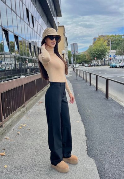 Suéter de cuello alto beige de manga larga, pantalones amplios negros, zapatos de plataforma color beige claro y un sombrero tipo cubo color beige claro. La modelo está de pie frente a un fondo de calle.