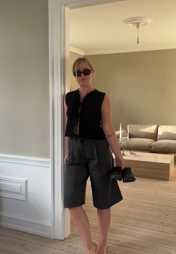 Zwarte cropped top met een strik aan de voorkant, donkergrijze geplooide shorts en zwarte schoenen in de hand. Minimalistisch interieur met houten vloeren.