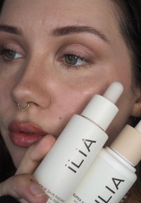 Twee ILIA huidverzorgingsflessen met witte en beige verpakkingen, met een minimalistisch ontwerp en opvallende etiketten. De achtergrond bevat een close-up van een gezicht.