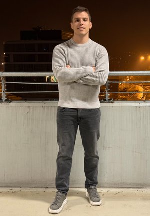 Suéter gris claro con textura acanalada, jeans oscuros y zapatillas grises. Persona de pie con los brazos cruzados frente a una pared texturizada durante la noche.