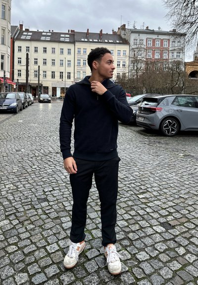 Sudadera negra con cremallera en el cuello, pantalones negros y zapatillas blancas con acentos naranjas. Calle de adoquines y fondo de edificio gris.