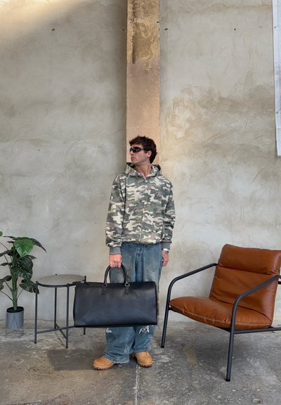 Hombre con sudadera con capucha camuflada, gafas de sol y vaqueros sosteniendo una bolsa negra de viaje, de pie entre una pequeña mesa con planta y una silla de cuero marrón en el interior.