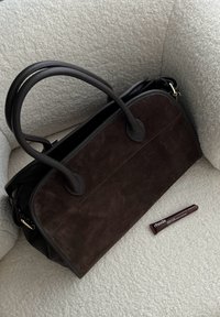 Braune Wildleder-Handtasche mit schwarzen Lederakzenten, ausgestattet mit zwei geschwungenen Griffen und einem Reißverschluss. Ein dunkles Kosmetiktube liegt daneben.