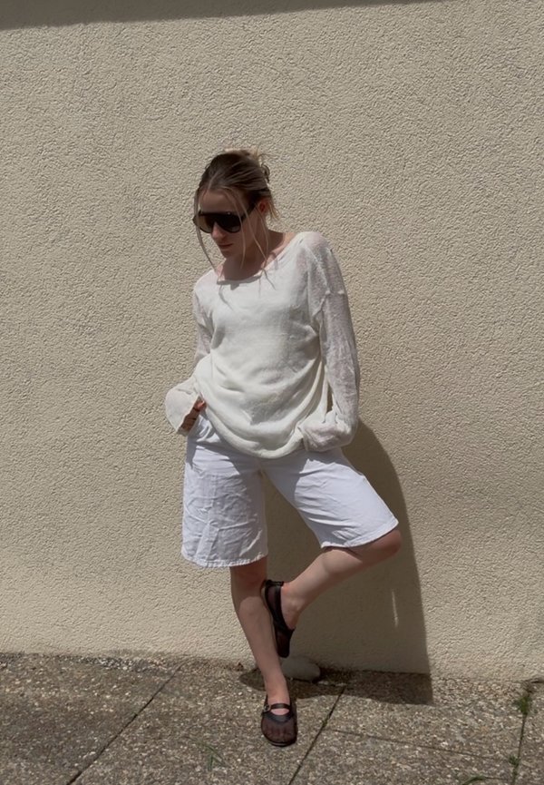 Femme portant un haut blanc à manches longues et un short, debout contre un mur beige texturé, mains dans les poches, une jambe pliée, portant des lunettes foncées et des chaussures.