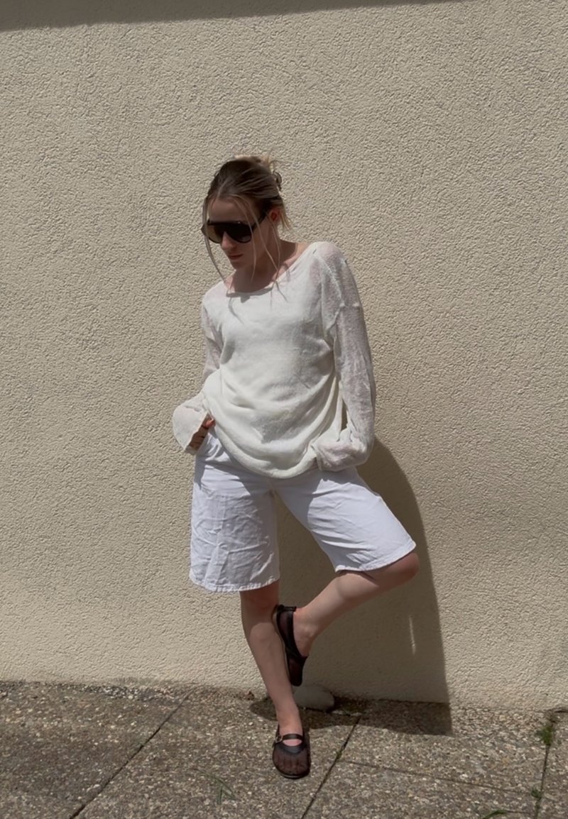 Femme portant un haut blanc à manches longues et un short, debout contre un mur beige texturé, mains dans les poches, une jambe pliée, portant des lunettes foncées et des chaussures.