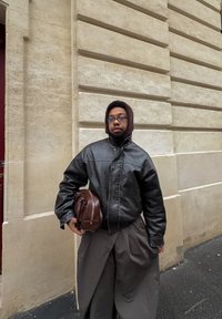 Mustast nahast jakk, hallid laiad püksid, hoides pruuni nahast kotti, seisab heledat värvi tekstuurse seina taustal.