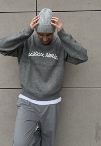 Grauer Strickpullover mit weiten Ärmeln und weißer, bestickter Schrift. Kombiniert mit grauer Hose und einer hellgrauen, gerippten Mütze.