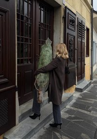 Une personne portant un long manteau sombre transporte un grand sapin de Noël vert avec une base marron, debout sur des marches en pierre devant des portes en bois.