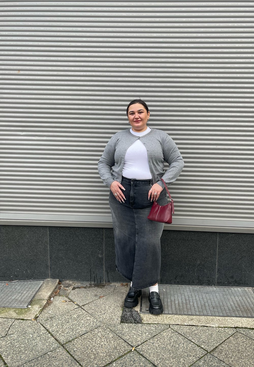 Cardigan gri peste un top alb, asociat cu o fustă lungă din denim negru. Pantofi cu platformă negri și o geantă mică roșie. Fundal texturat.
