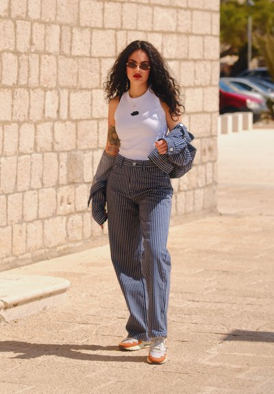 Pantalones de pata de elefante a rayas azul y blancas combinados con un top sin mangas blanco. Una camisa con estampado colgada del brazo y gafas de sol completan el look.